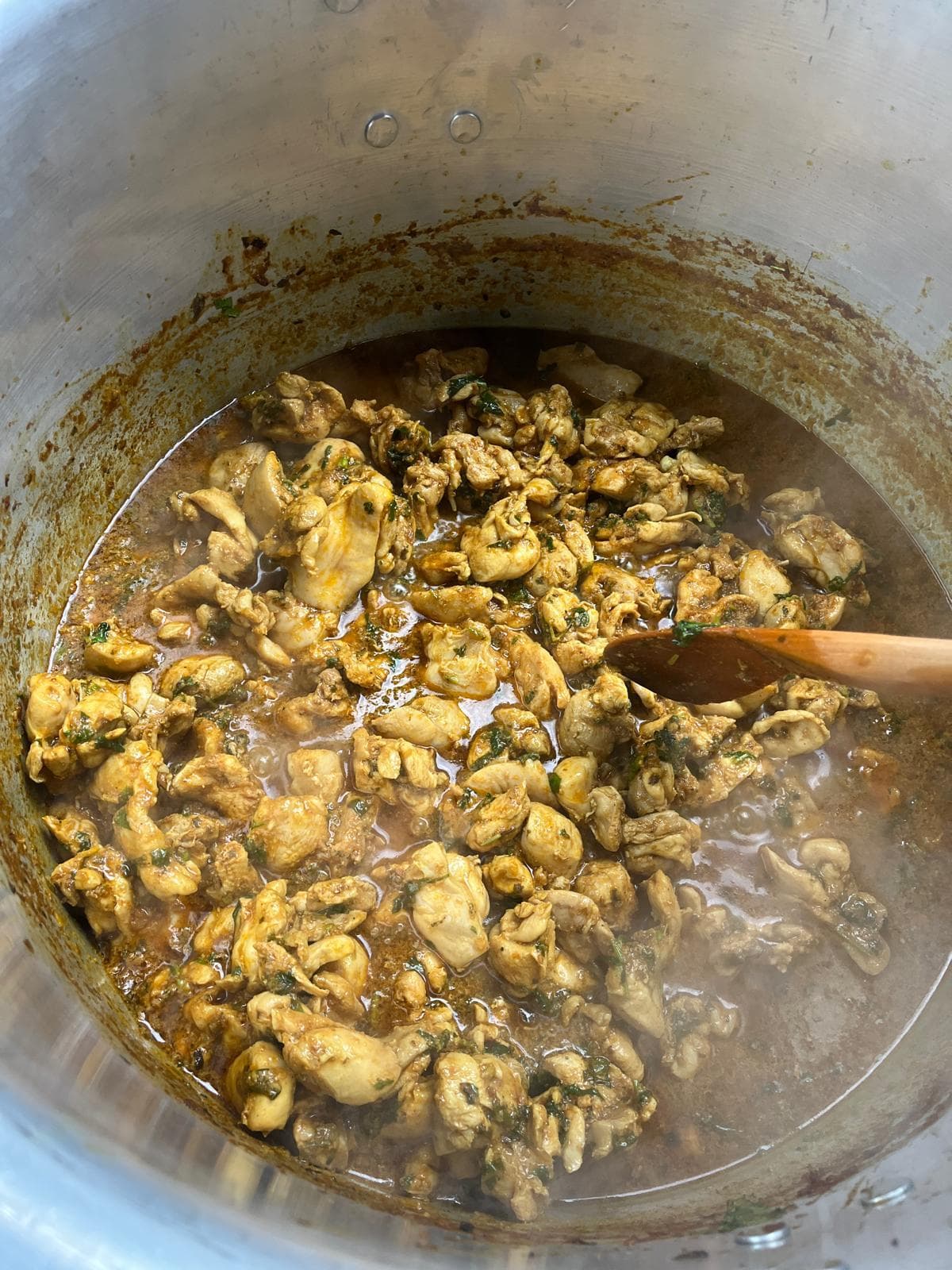 Chicken and coriander simmering in a large aluminium karoga pot, part-stirred with a long wooden spoon.