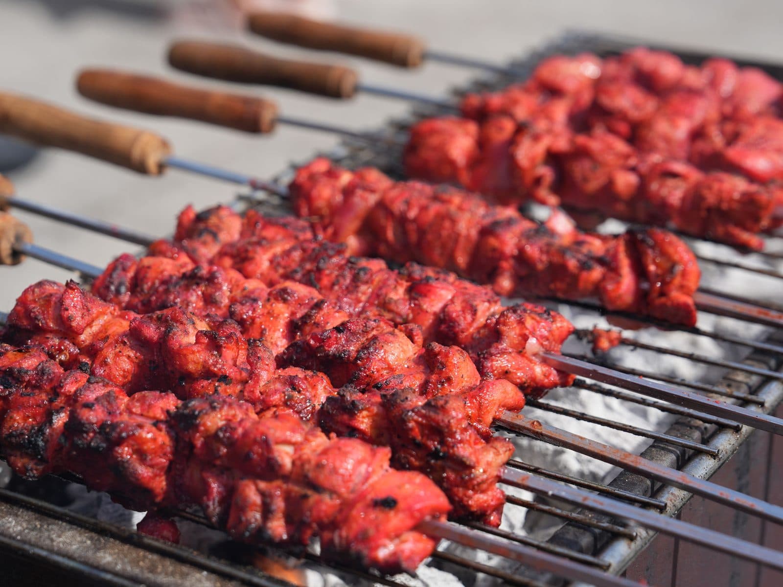 A row of wooden-handled skewers loaded with bright red marinated chicken, charring over an open charcoal grill.
