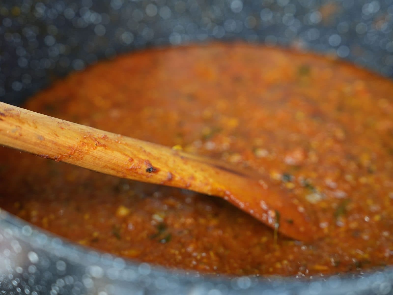 A wide pot of deep orange curry sauce flecked with spices and coriander, part-stirred with a long wooden spoon.