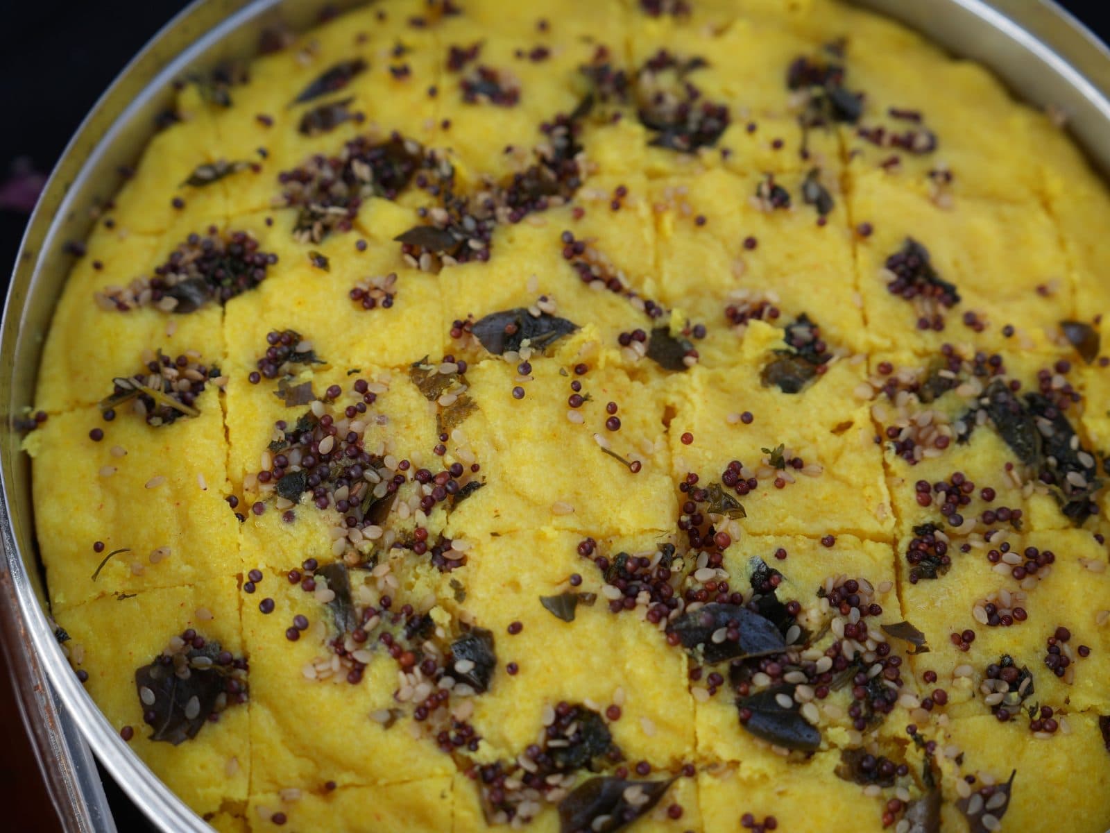 A wide steel tray of yellow dhokla fresh from the steamer, scored into diamonds and topped with black mustard seeds and curry leaves.