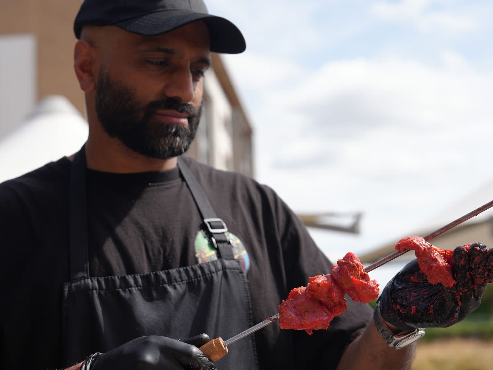 A cook in a black cap, apron and gloves sliding charred marinated meat off a long metal skewer with tongs, under an open sky.