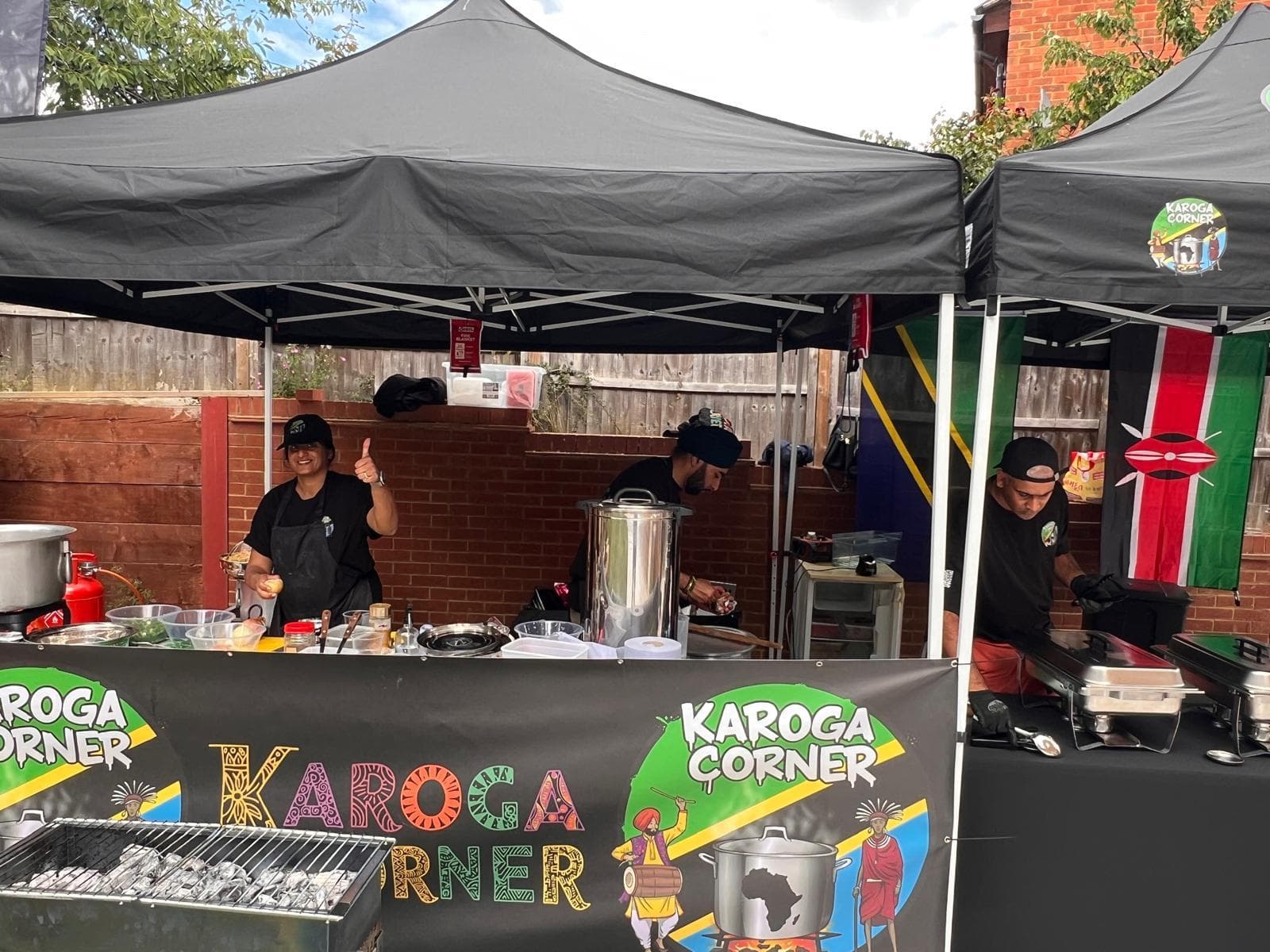 Three Karoga Corner cooks working under joined gazebos at a private event, with Kenyan and Tanzanian flags hung behind them and a charcoal grill loaded at the front.