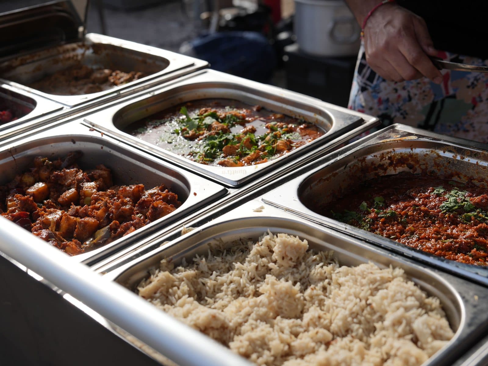 Four steel serving trays along the hot counter — pilau rice at the front, a dark potato masala, a curry finished with coriander and a deep red sauce behind — with a member of the team serving from the far side.