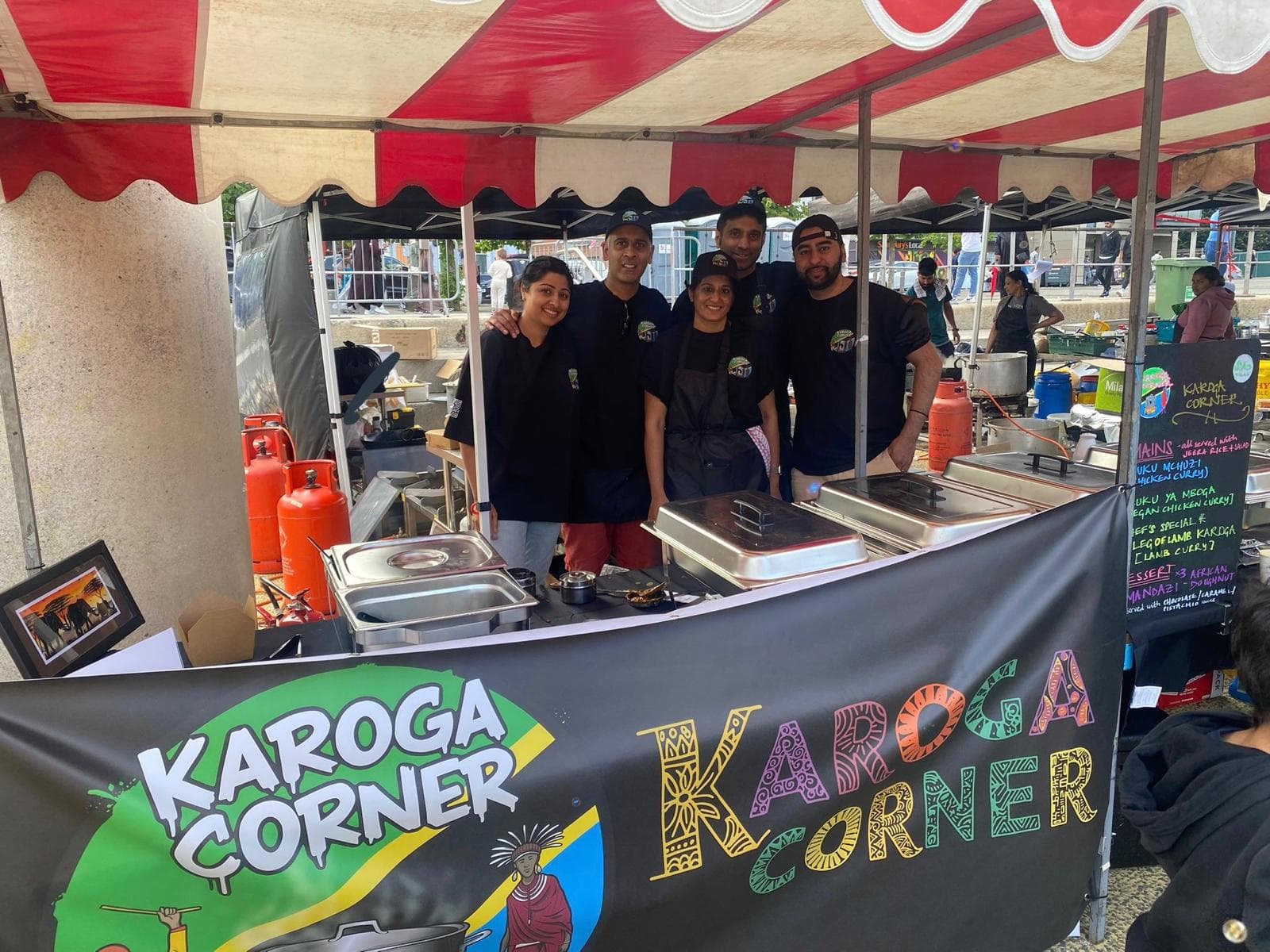 Five of the Karoga Corner team in branded shirts behind their counter at a street market, with the day's chalkboard menu of kuku mchuzi, kuku ya mboga and leg of lamb karoga alongside.