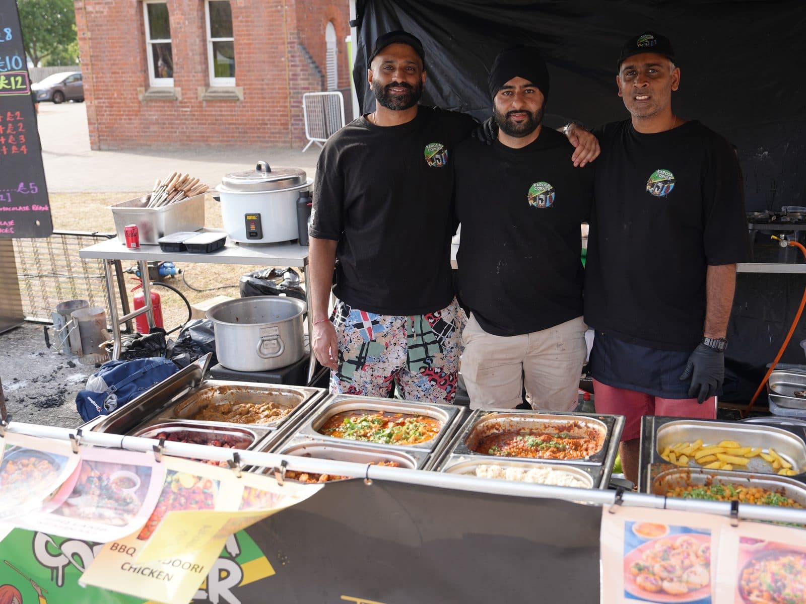 The full Karoga Corner counter at a street market: three of the team behind a run of serving trays, rice cookers and menu boards, with a red brick building behind.