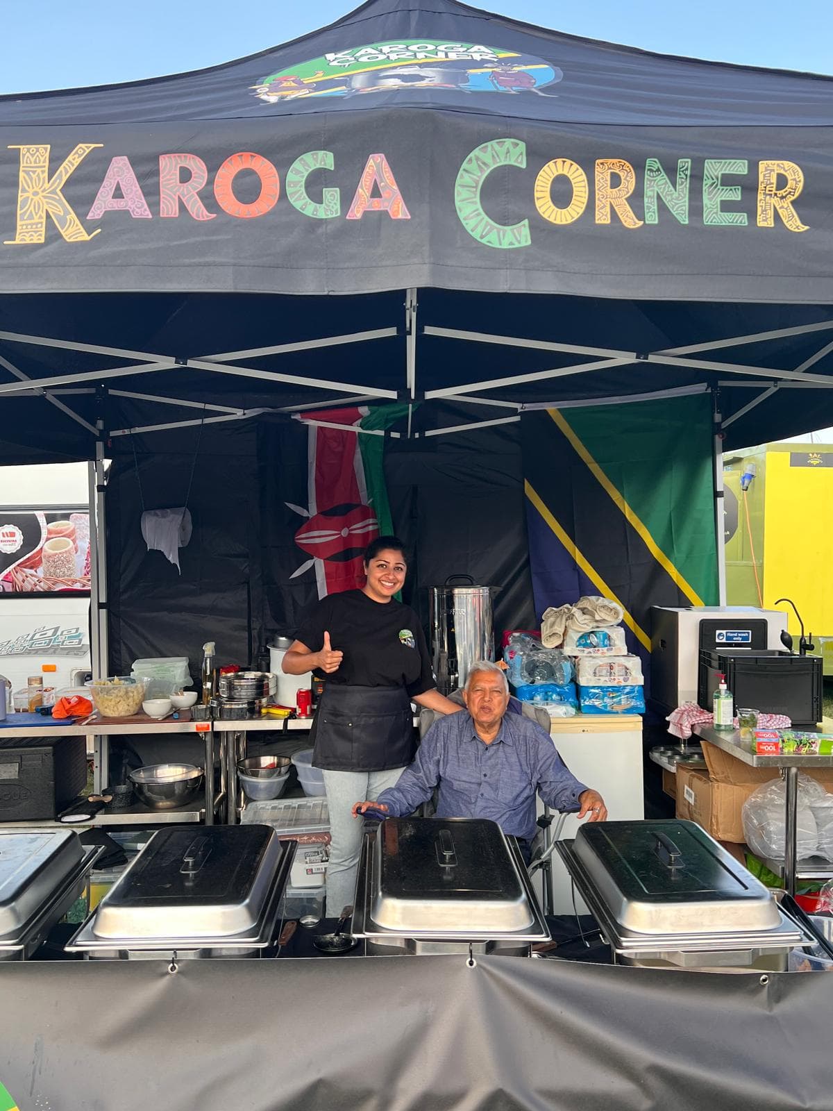 Two members of the Karoga Corner team under the stall's black gazebo, with the hand-painted Karoga Corner sign above them and Kenyan and Tanzanian flags behind.